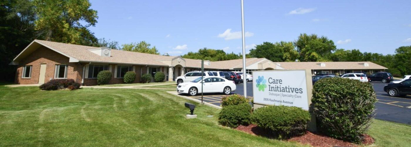 A single-story care facility with a brick exterior is surrounded by a well-maintained lawn. An American flag on a flagpole stands in front. Nearby, a sign reads Care Initiative, and several cars are parked in the lot under a clear blue sky. The brand name of the facility is Care Initiative.