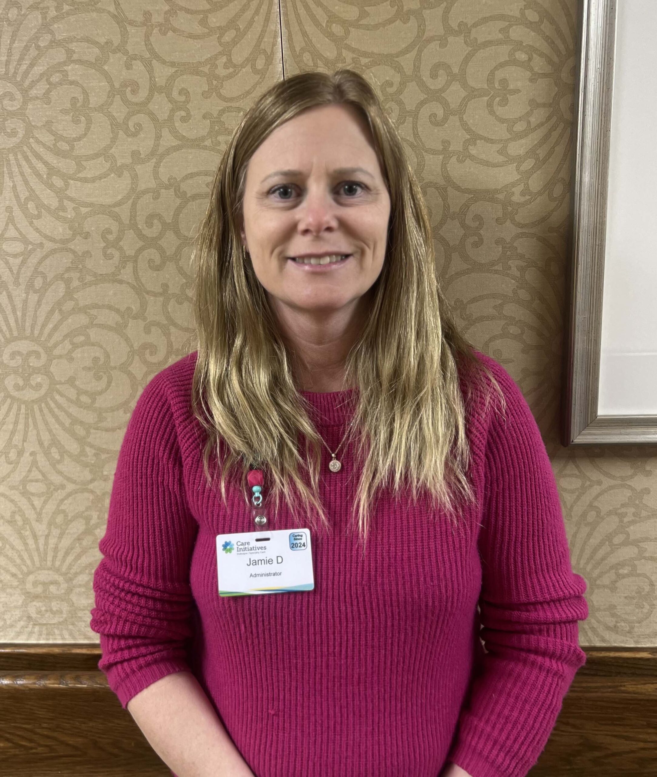 A person with long hair in a pink sweater stands before a patterned wall, showcasing their name badge as part of the Care Initiative.