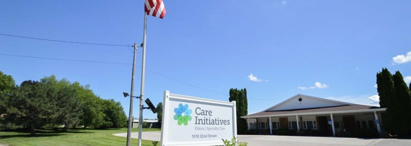 A sunny day at a Care Initiative facility showcases a well-maintained lawn and lush trees. The American flag waves proudly beside the Care Initiative sign, while a driveway gracefully leads to a building with an inviting portico.