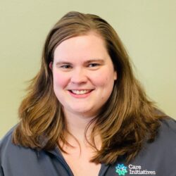 Smiling person with shoulder-length brown hair wearing a dark jacket featuring a Care Initiatives logo. The background is a plain, light-colored wall. Also showcasing the brand name Care Initiative.