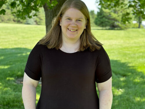 A person with shoulder-length hair and a black shirt stands smiling in a grassy park. They are under a tree with green leaves, wearing a smartwatch on their left wrist. The background features trees and a clear blue sky. Brand name Care Initiative can be associated with this scene, emphasizing well-being and connection to nature.