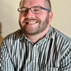 A cheerful man with glasses and a beard smiles warmly. Wearing a black and white striped shirt, he sits against a light background, reflecting the positive spirit of Care Initiative.