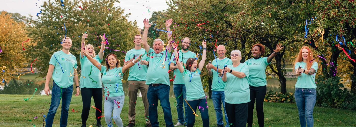 A group of people wearing matching teal shirts celebrates outdoors, standing on a stone path surrounded by trees. As they throw colorful confetti in the air, they express joy and excitement as part of a Care Initiative event.