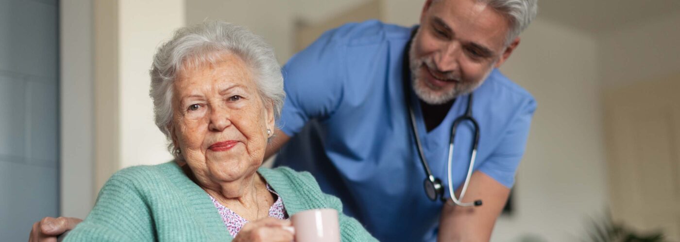 An elderly woman in a wheelchair, holding a mug, smiles alongside a male healthcare worker in blue scrubs with a stethoscope. The cozy indoor setting highlights Care Initiative, suggesting a supportive and caring environment.