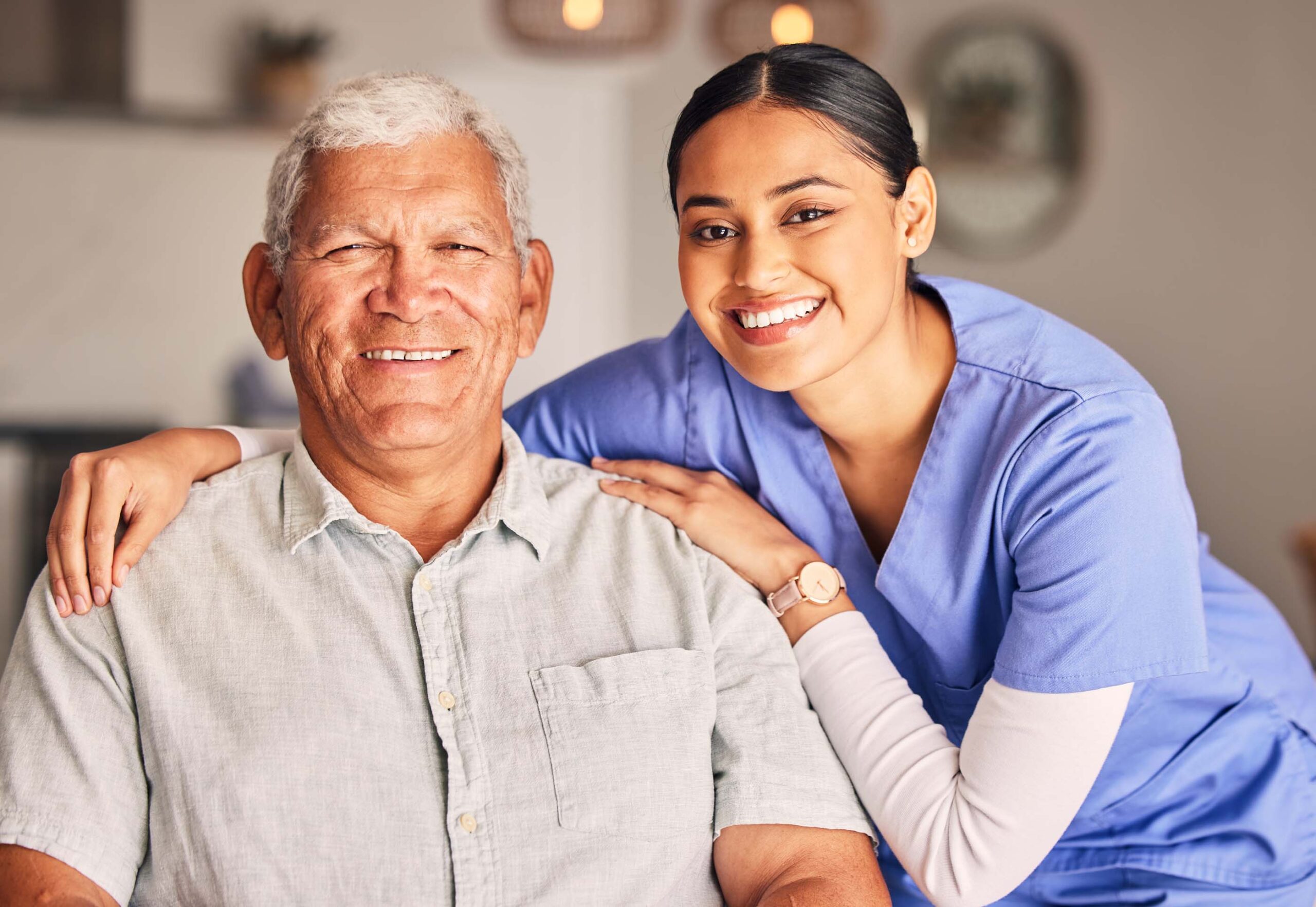 A smiling elderly man in a light gray shirt sits as a young woman in blue scrubs rests her arms on his shoulders, embodying the essence of Care Initiative. They are in a bright room, radiating warmth and companionship.