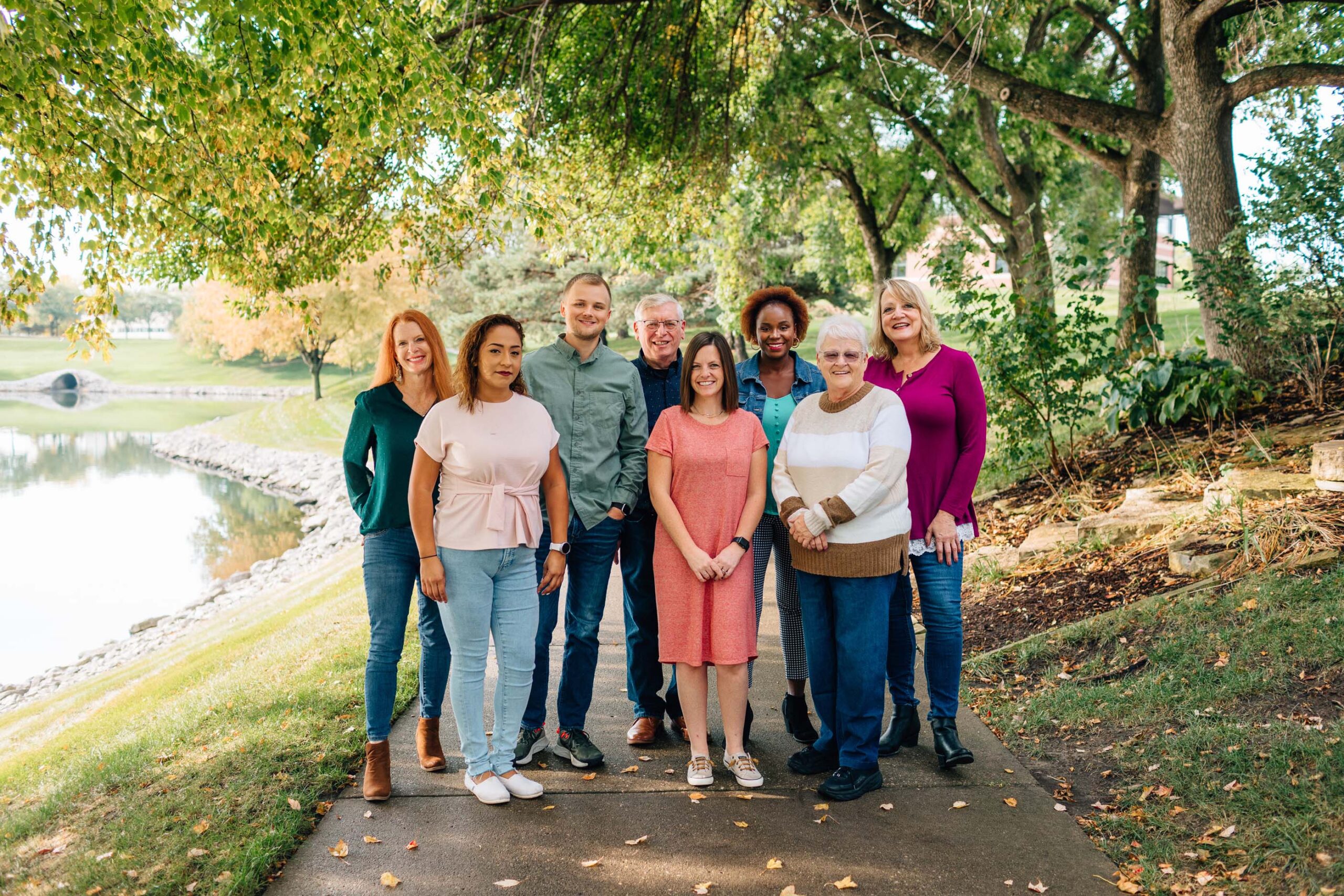 A group of eight people stands on a pathway surrounded by trees and a pond, smiling at the camera. Part of Care Initiative, they are dressed casually in various outfits, with autumn leaves scattered on the ground and in the trees.