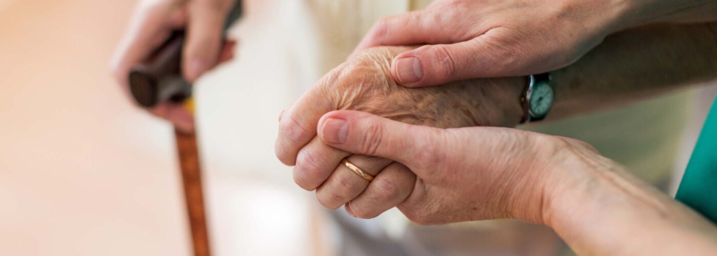 A close-up of a caregiver gently holding the hand of an elderly person as the elderly person grips a walking cane, symbolizing support and compassion. Care Initiative.