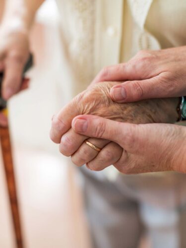 A close-up of a caregiver gently holding the hand of an elderly person as the elderly person grips a walking cane, symbolizing support and compassion. Care Initiative.