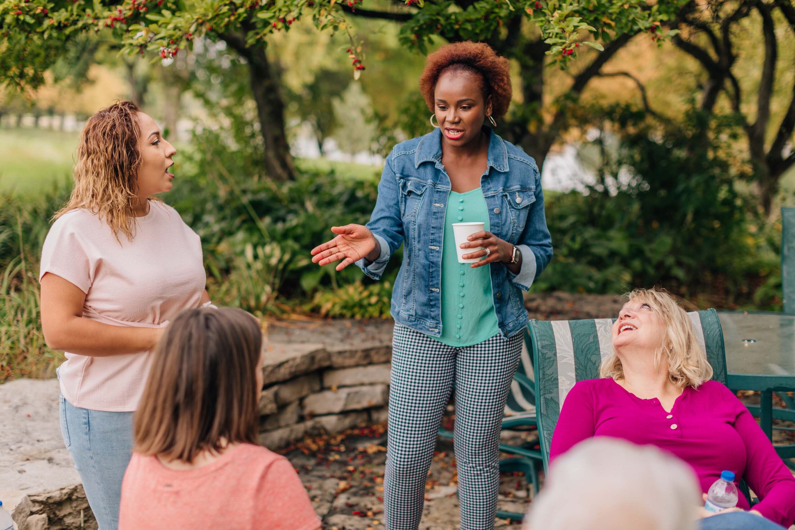 A group of women are gathered outdoors, talking and smiling. One woman holding a cup speaks while the others listen. Trees and greenery create a serene backdrop, suggesting a friendly atmosphere that reflects their involvement in the Care Initiative.