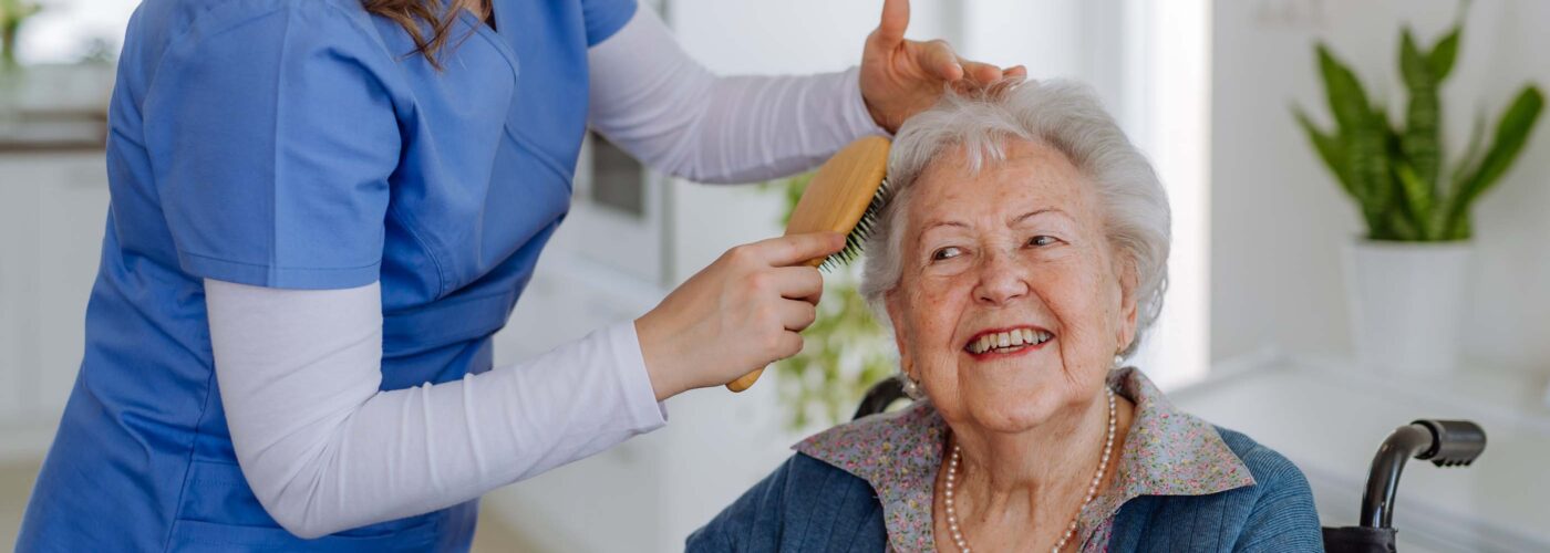 A caregiver in a blue uniform gently brushes the hair of a smiling elderly woman in a wheelchair. They are in a bright, well-lit room with a plant visible in the background, highlighting the Care Initiative they follow.