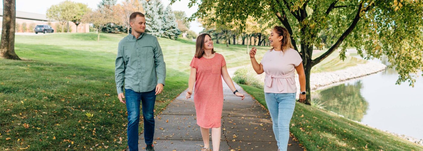 Three people stroll along a tree-lined path. Two women, one in a pink dress and the other in a pink top and jeans, converse happily as part of the Care Initiative. A man in a green shirt and jeans joins them. The path is framed by lush greenery and a calm waterway.