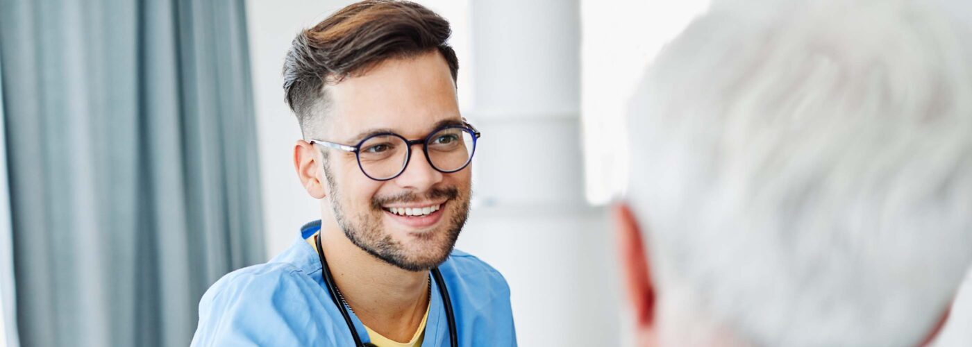 A healthcare professional in blue scrubs, smiling and wearing glasses, warmly holds the shoulder of an older person with gray hair. The setting is indoors, featuring a curtain and a white pillar in the background, highlighting a compassionate Care Initiative atmosphere.