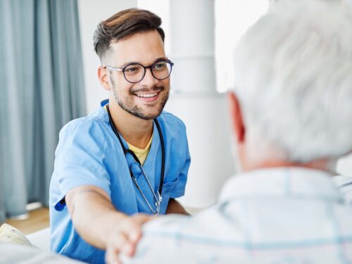 A healthcare professional in blue scrubs, smiling and wearing glasses, warmly holds the shoulder of an older person with gray hair. The setting is indoors, featuring a curtain and a white pillar in the background, highlighting a compassionate Care Initiative atmosphere.