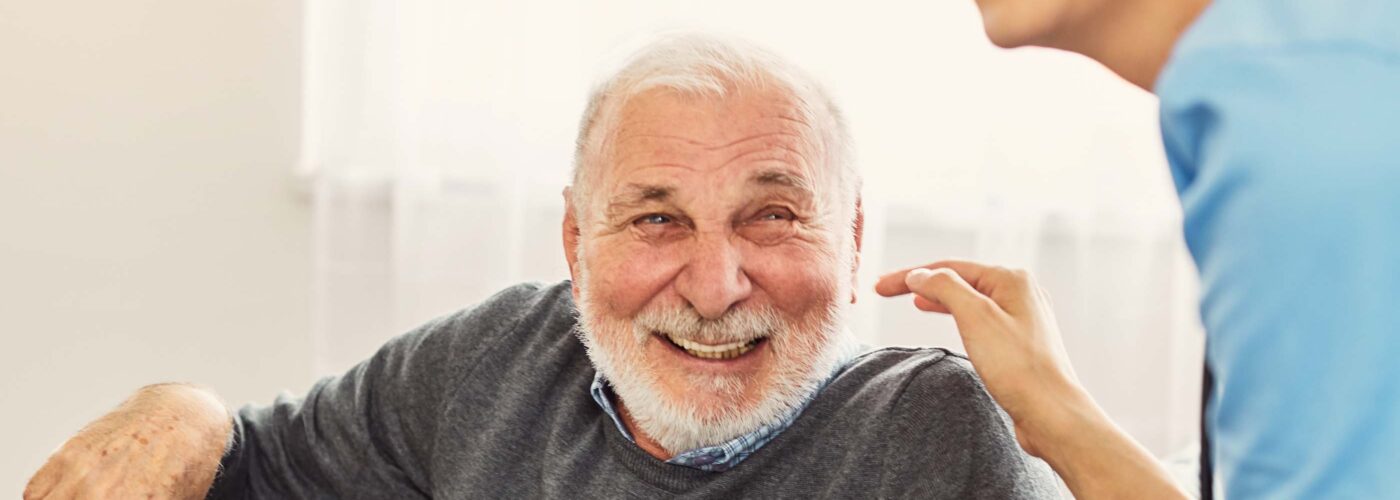 An elderly man with a white beard sits on a couch, smiling and holding a walking cane. A cheerful woman in a blue shirt is next to him, holding his hand. They both seem to be laughing and enjoying each other's company in a well-lit room, highlighting the success of Care Initiative.