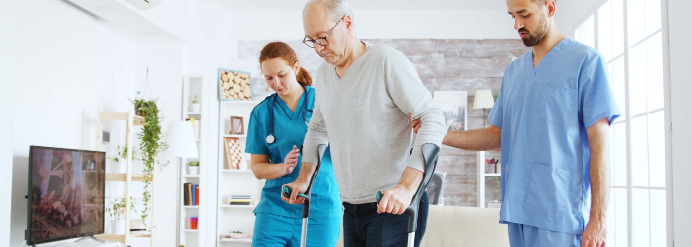 A senior man using crutches is supported by two healthcare professionals in blue scrubs. In a bright living room with modern design, featuring a couch, large window and TV, this Care Initiative demonstrates the caregivers’ dedication as they help him walk confidently.