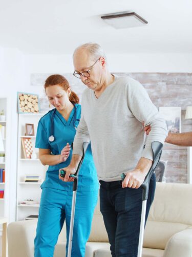 A senior man using crutches is supported by two healthcare professionals in blue scrubs. In a bright living room with modern design, featuring a couch, large window and TV, this Care Initiative demonstrates the caregivers’ dedication as they help him walk confidently.