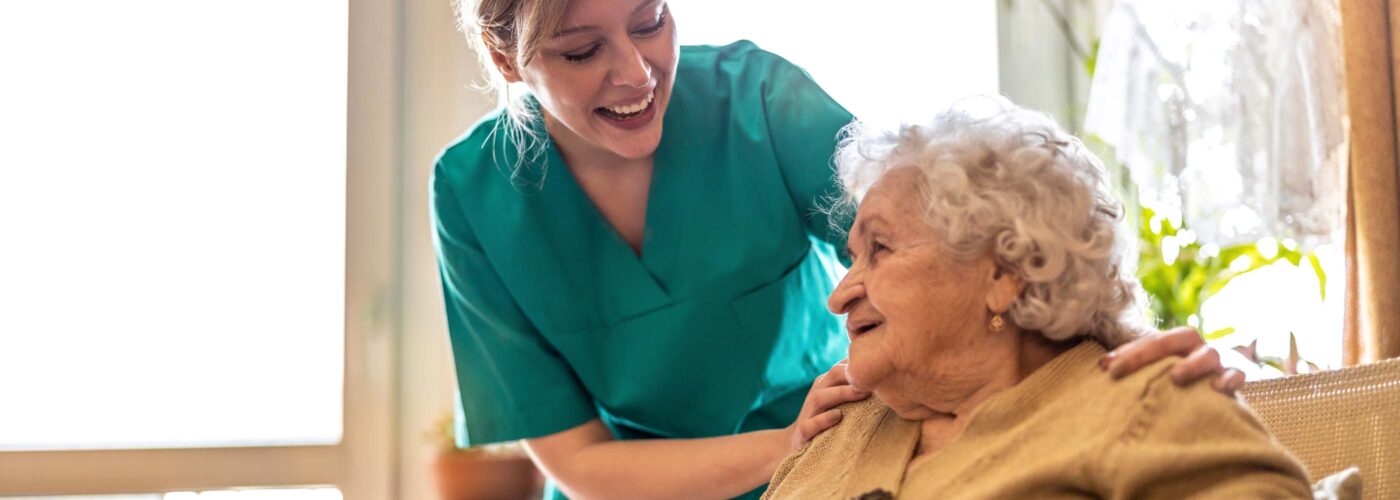 A smiling caregiver in a teal uniform gently places her hand on the shoulder of an elderly woman with white hair, as they both share a warm smile. They are indoors with natural light streaming through a window and vibrant green plants in the background, embodying a nurturing care initiative by Care Initiative.