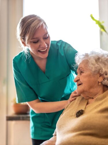 A smiling caregiver in a teal uniform gently places her hand on the shoulder of an elderly woman with white hair, as they both share a warm smile. They are indoors with natural light streaming through a window and vibrant green plants in the background, embodying a nurturing care initiative by Care Initiative.