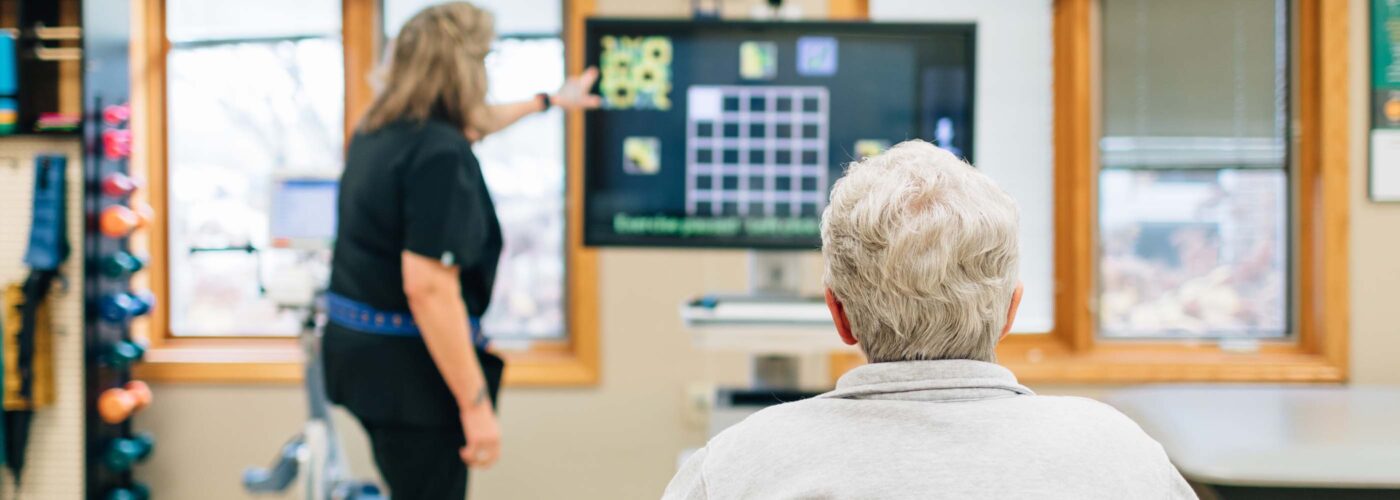 A person in a wheelchair observes as a staff member points at a monitor displaying a grid with images and the text Word Search. The setting, part of the Care Initiative program, resembles a therapy room with exercise equipment and large windows in the background.