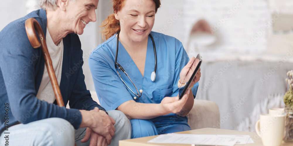 A nurse in blue scrubs shows something on a tablet to an elderly man seated next to her. Holding a cane, he smiles warmly as they discuss the Care Initiative in the cozy room with a table in front of them.