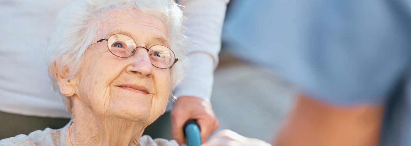 An elderly woman with glasses and white hair sits in a wheelchair, smiling warmly at a healthcare worker who is holding her hands. She wears a light gray sweater, set against a softly blurred background, embodying the compassion of Care Initiative.