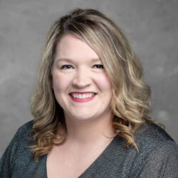 A person with shoulder-length blonde hair is smiling at the camera, wearing a dark textured top. The gray neutral background enhances a sense of warmth and approachability, reflecting a spirit of care. The brand name Care Initiative embodies this welcoming presence.