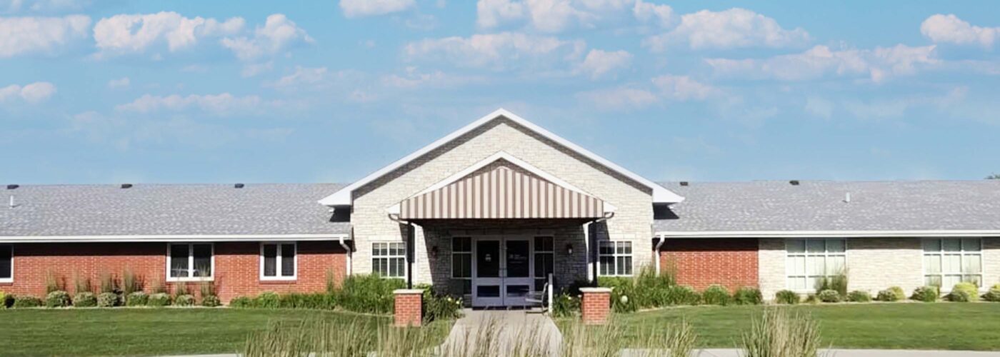 A single-story brick building with a striped awning over the entrance is surrounded by neatly trimmed grass and shrubs under a bright blue sky. This Care Initiative location blends seamlessly with its surroundings.