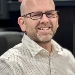 A man with glasses and a light-colored, patterned shirt smiles while sitting. The background is slightly blurred, featuring dark furniture. This warm scene radiates a sense of community and support, embodying the essence of the Care Initiative brand.