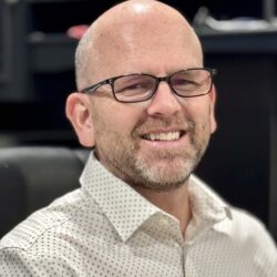 A man with glasses and a light-colored, patterned shirt smiles while sitting. The background is slightly blurred, featuring dark furniture. This warm scene radiates a sense of community and support, embodying the essence of the Care Initiative brand.