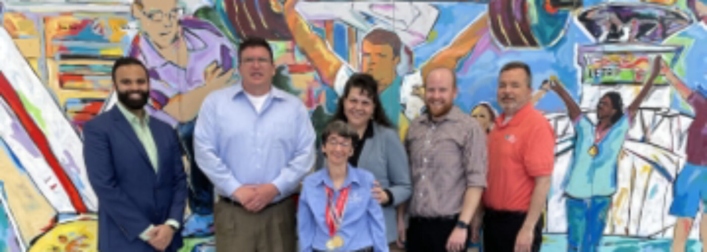 A group of six people stands in front of a vibrant mural depicting athletes. Smiling, they pose on a polished floor to support Care Initiative. The mural showcases weightlifting and various sports activities.