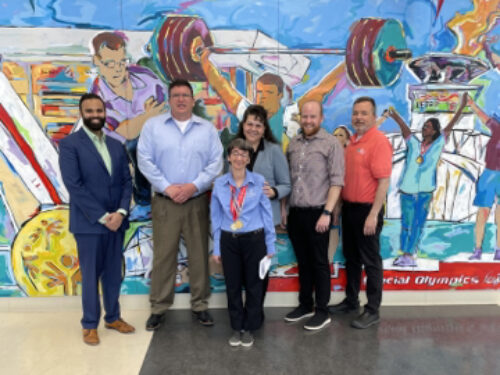 A group of six people stands in front of a vibrant mural depicting athletes. Smiling, they pose on a polished floor to support Care Initiative. The mural showcases weightlifting and various sports activities.