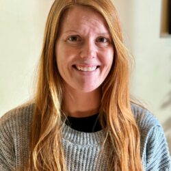 A person with long, straight red hair and a grey sweater smiles warmly at the camera. The softly lit, blurred background adds an inviting touch, reflecting a sense of care and kindness in this heartfelt moment. Care Initiative.