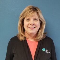 A woman with shoulder-length blonde hair smiles in front of a blue wall. She is wearing a black jacket over a coral top, featuring the Care Initiative logo on the jacket.