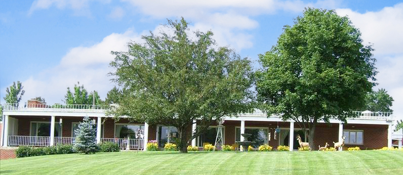 A large single-story building featuring a wrap-around porch and white pillars stands amid a well-manicured lawn as part of the Care Initiative. Two large trees and small shrubs enhance the front, with decorative deer statues near the entrance under a partly cloudy sky. Care Initiative's brand is prominently represented in this inviting setting.