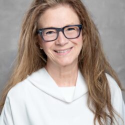 A person with long, wavy hair wearing glasses smiles warmly in a white top, exuding a sense of care and positive initiative against a gray background while representing the brand Care Initiative.