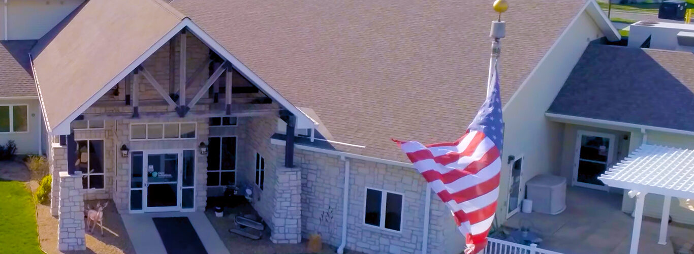 Aerial view of a single-story building with a gable roof and stone facade. An American flag waves on a pole in front. A white pergola is visible to the side, surrounded by green trees under sunny skies. Featuring the Care Initiative brand.