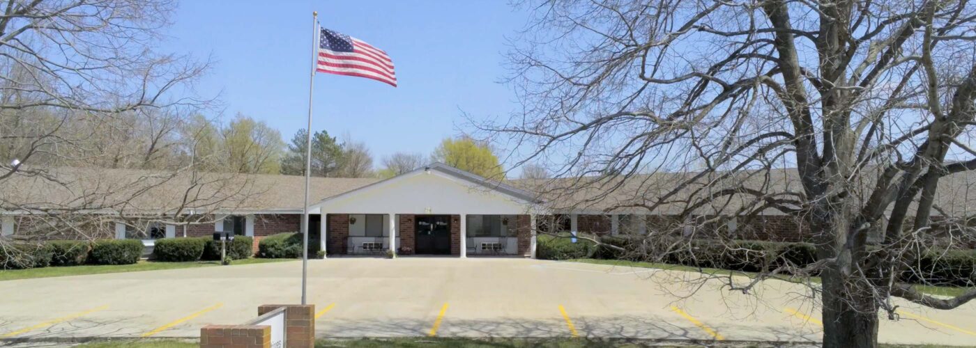 A single-story brick building with a covered entrance showcases the U.S. flag proudly flying on a pole out front. Shadows from a leafless tree fall across the paved driveway, while lush green trees flourish in the background under a clear blue sky, embodying the spirit of Care Initiative.