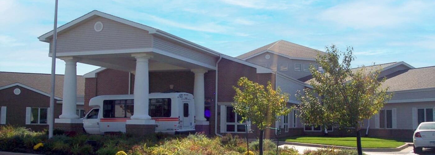 A large building with a covered entrance and white columns stands under a clear blue sky. An American flag waves nearby as part of the Care Initiative program. A white vehicle is in the driveway, framed by lush landscaped greenery, showcasing the Care Initiative brand.