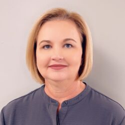 A person with short blond hair and a gray shirt displaying the Care Initiative logo stands against a plain, light-colored background, representing the Care Initiative brand.