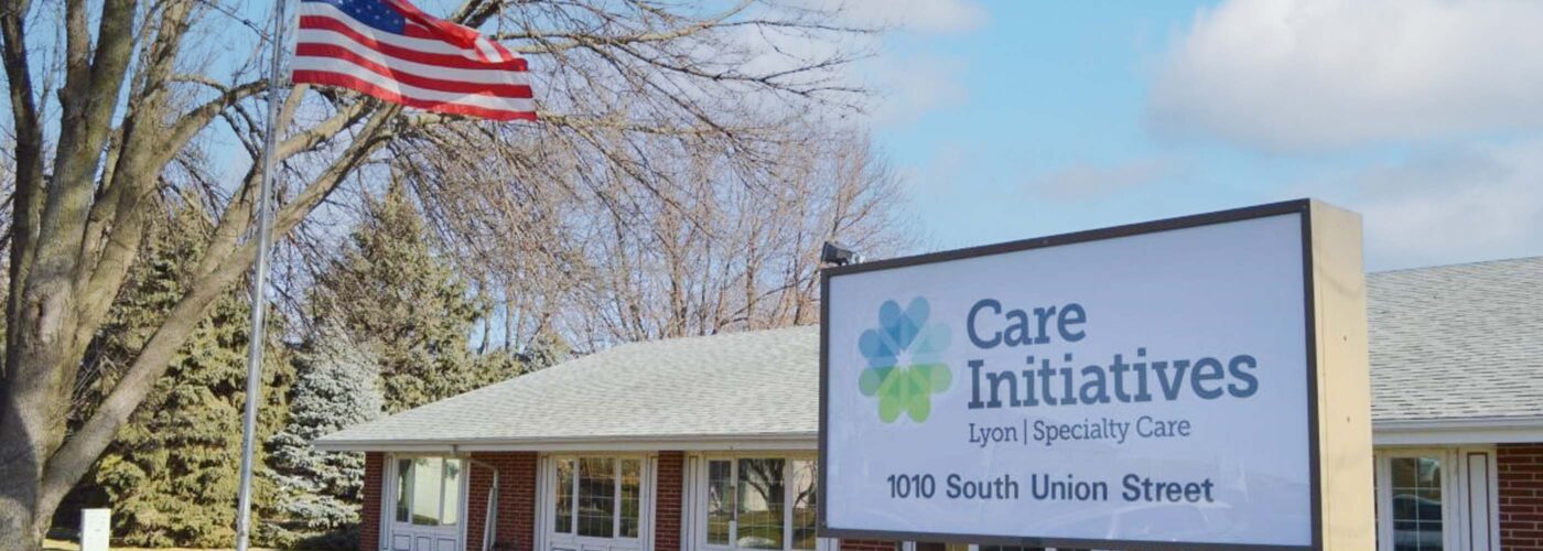 An American flag waves near a brick building with a sign for Care Initiative Lyon Specialty Care at 1010 South Union Street. The scene includes trees and a partly cloudy sky, along with the brand name Care Initiative prominently displayed.