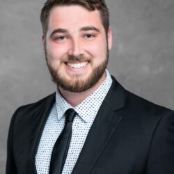 A man with short hair and a beard smiles at the camera. He wears a black suit, white patterned shirt, and black tie against a plain gray backdrop, depicting a modern image that could be part of an awareness campaign by Care Initiative.