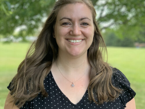 A woman with long brown hair smiles at the camera, wearing a black top with white polka dots and a silver necklace. The background features a green, grassy area and trees. Care Initiative.