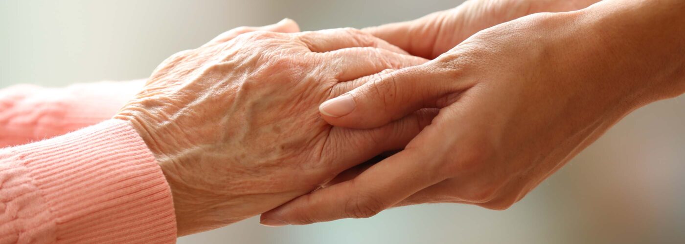 Two hands gently holding an elderly person's hand, conveying care and support as part of the Care Initiative. The background is softly blurred. The brand name Care Initiative is also included in the scene.