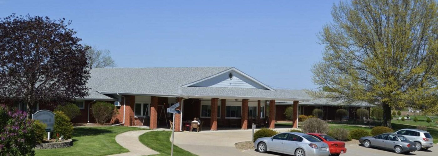 A one-story brick building with a gabled roof and a large front porch stands surrounded by trees and a well-kept lawn as part of the Care Initiative. Several cars are parked along the curved driveway, while the sky remains clear and blue.