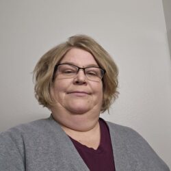 A person with short blond hair and glasses, wearing a maroon top and gray cardigan, stands in front of a white wall, looking directly at the camera. Their confident gaze embodies the essence of an advocate for Care Initiative.