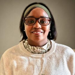 A person wearing glasses and a headband smiles at the camera, dressed in a light-colored sweater with a patterned shirt underneath, accessorized with a necklace and earrings. The background is plain and softly lit. Care Initiatives.