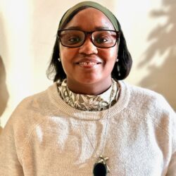 A person wearing glasses, a green headscarf, and a light-colored sweater stands against a bright background. With a warm smile, they proudly display their necklace featuring a large oval pendant, embodying the essence of care. Care Initiative.