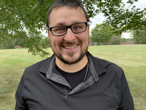 A person with short dark hair and glasses is smiling while standing outdoors. They are wearing a black shirt with a gray collar and a necklace. Behind them is a grassy field with trees and a cloudy sky. Care Initiative.