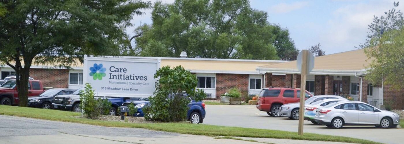 A brick building with a sign reading Care Initiative is surrounded by trees. Several cars are parked on the paved driveway in front of the facility, under a partly cloudy sky. Care Initiative.