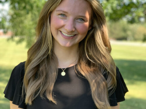 A woman with long, light brown hair smiles at the camera. She is wearing a black top and standing outdoors in a grassy area with trees and sunlight in the background. Care Initiatives.